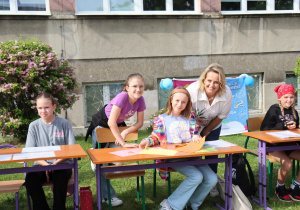Na zdjęciu pięć dziewczynek i nauczycielka uczestniczą w plenerowych zajęciach przy stolikach ustawionych na trawie. Uczennice rysują i kolorują kartki papieru, a nauczycielka pochyla się i uśmiecha do kamery razem z jedną z dziewczynek. W tle znajduje się budynek szkoły, krzew z różowymi kwiatami oraz kolorowy baner i niebieskie balony. Atmosfera jest pogodna, twórcza i pełna zaangażowania.
