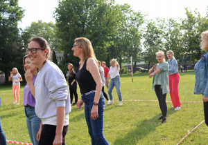 Grupa dorosłych kobiet bierze udział w zabawie lub rozgrzewce na trawiastym boisku. Uczestniczki uśmiechają się, rozmawiają i wykonują ruchy rękami, stojąc w wyznaczonym kręgu oznaczonym czerwoną taśmą. W tle widoczne są drzewa oraz osoby spacerujące po boisku. Panuje słoneczna pogoda i radosna atmosfera wspólnej aktywności.