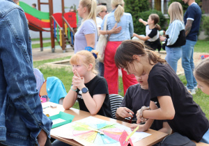 Na zdjęciu kilkoro dzieci siedzi i stoi przy stole ustawionym na świeżym powietrzu, skupiając się na grze z użyciem kolorowego koła fortuny wykonanego z papieru. Jedna z dziewczynek pochyla się, wskazując coś na planszy, a pozostałe dzieci obserwują. W tle widoczne są osoby dorosłe i dzieci, plac zabaw z kolorową zjeżdżalnią oraz trawiasty teren. Atmosfera jest aktywna i zaangażowana, typowa dla festynu szkolnego.