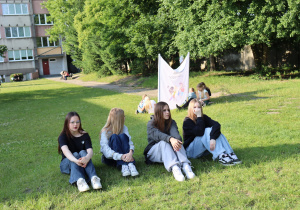 Na zdjęciu cztery nastolatki siedzą na trawie w cieniu drzew, odpoczywając podczas szkolnego pikniku. Wszystkie dziewczyny mają swobodny, sportowy ubiór, a ich postawa wskazuje na relaks i rozmowę. W tle, pod drzewami, widoczne są inne osoby – niektóre siedzą, inne spacerują. Po prawej stronie rozciągnięto biały baner z ilustracjami dzieci i napisem. W oddali widać budynek szkolny. Scena ukazuje spokojny moment wydarzenia na świeżym powietrzu.