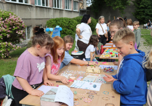 Na zdjęciu dzieci stoją przy stołach ustawionych na zewnątrz szkoły, prezentując ręcznie wykonane ozdoby i biżuterię, takie jak bransoletki, naszyjniki, naklejki i breloczki. Dzieci są zaangażowane, rozmawiają i pokazują przedmioty odwiedzającym. W tle widoczne są inne stoiska, uczniowie, rodzice i nauczyciele uczestniczący w wydarzeniu, a także krzewy i budynek szkoły. Atmosfera jest kolorowa, radosna i twórcza.