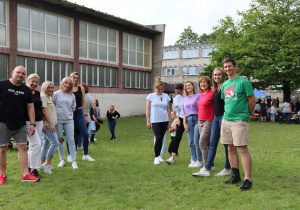 Na zdjęciu grupa dorosłych – nauczycieli i rodziców – stoi w rzędzie na trawie przed budynkiem szkolnej sali gimnastycznej. Wszyscy uśmiechają się i patrzą w stronę aparatu, pozując do wspólnego zdjęcia. W tle widać uczestników pikniku, dzieci, kolorowe namioty i zjeżdżalnie, a także drzewa i budynki szkolne. Atmosfera jest pogodna, integracyjna i radosna.