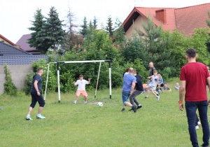 Na zdjęciu rozgrywany jest mecz piłki nożnej na trawiastym boisku. Kilku dorosłych i dzieci biegnie w stronę bramki, w której stoi młody bramkarz w białej koszulce i różowych spodenkach, gotowy do obrony strzału. Piłka znajduje się tuż przed linią bramkową. W tle widać widzów siedzących na trawie oraz domy i drzewa otaczające boisko. Scena przedstawia dynamiczną i radosną integrację międzypokoleniową podczas szkolnego pikniku.
