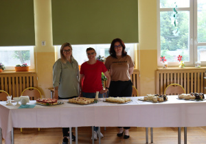 Na zdjęciu trzy kobiety stoją za stołem nakrytym białym obrusem w szkolnej sali. Na stole ustawiono kilka tac z pokrojonymi kawałkami ciast, w tym ciasta z owocami i ciasta z polewą czekoladową. Za kobietami widać okna z opuszczonymi roletami, ozdobione doniczkami z kwiatami i papierowymi dekoracjami. Atmosfera jest serdeczna i gościnna, typowa dla kawiarenki organizowanej podczas szkolnych wydarzeń.