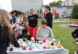 Na zdjęciu uczennica siedzi za stołem przykrytym obrusem, na którym rozłożone są kolorowe, ręcznie wykonane maskotki szydełkowe w kształcie zwierzątek i jedzenia. Przed stoiskiem stoi kobieta i inna uczennica oglądająca produkty. W tle widać plac zabaw, budynek szkoły oraz tłum uczestników pikniku, którzy spacerują i odwiedzają kolejne stoiska. Scena ukazuje twórczy charakter wydarzenia i aktywność uczniów.