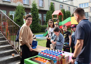 Na zdjęciu kobieta i mężczyzna obsługują stoisko z napojami i przekąskami na terenie szkolnego placu zabaw. Na stole znajdują się rzędy butelek z sokami i wodą oraz paczki chipsów. Przed stoiskiem stoi grupa dzieci, z których jedno przelicza pieniądze, przygotowując się do zakupu. W tle widoczny jest kolorowy plac zabaw oraz budynek szkoły. Atmosfera jest pogodna i towarzyska, typowa dla wydarzenia plenerowego.