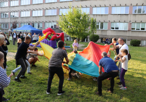 Na zdjęciu grupa dzieci i dorosłych uczestniczy w zabawie z użyciem dużej, kolorowej chusty animacyjnej na trawiastym terenie przed szkołą. Chusta jest uniesiona i poruszana energicznie, a część osób znajduje się pod nią. W tle widoczne są dmuchana zjeżdżalnia, niebieski namiot z napisem „Park Trampolin Air Jump”, drzewa oraz budynek szkolny z licznymi oknami. Atmosfera jest dynamiczna, radosna i integracyjna.