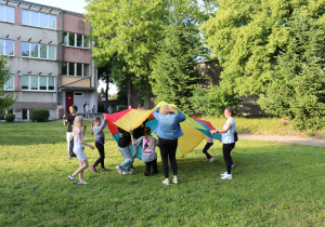 Na zdjęciu grupa dzieci oraz kilka dorosłych osób trzyma kolorową chustę animacyjną w barwach czerwieni, żółci, zieleni i niebieskiego. Dzieci unoszą ją do góry, tworząc z niej kolorowy namiot. Zabawa odbywa się na trawiastym terenie przed budynkiem szkoły, otoczonym wysokimi drzewami. W tle widać inne osoby spacerujące oraz szkolny budynek z dużymi oknami. Atmosfera jest pogodna, radosna i sprzyja integracji.