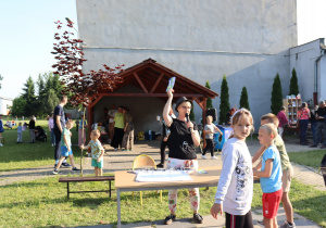 Na zdjęciu kobieta w okularach i kapeluszu trzyma mikrofon i kolorową nagrodę, stojąc przy stole podczas szkolnego pikniku. Prawdopodobnie prowadzi konkurs lub loterię. Przed nią stoją dzieci, a w tle widoczna jest wiata z grupą osób oraz inne atrakcje, m.in. regał z nagrodami i miejsce gastronomiczne. Po lewej stronie bawią się dzieci, a po prawej widać przechadzających się uczestników wydarzenia. Pogoda jest słoneczna, a atmosfera pełna radości i ruchu.