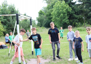 Na zdjęciu grupa chłopców stoi wokół bramki na trawiastym boisku. Część z nich rozmawia, inni patrzą w stronę aparatu. W tle widać innych uczestników wydarzenia, w tym osoby siedzące na krzesłach i dzieci w zielonych koszulkach. Otoczenie stanowią drzewa i wysoka trawa. Scena przedstawia moment odpoczynku lub zmiany drużyn podczas meczu w ramach szkolnego pikniku.