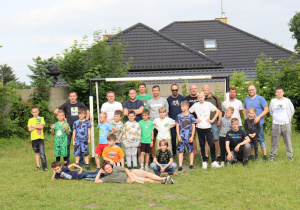 Na zdjęciu grupa dzieci i dorosłych pozuje wspólnie do pamiątkowego zdjęcia na boisku piłkarskim. W tle widoczna jest bramka oraz dom z ciemnym dachem otoczony zielenią. Uczestnicy – zarówno chłopcy, jak i mężczyźni – stoją i siedzą na trawie, uśmiechając się i patrząc w stronę aparatu. Część dzieci pozuje w strojach sportowych, co sugeruje, że uczestniczyli wcześniej w meczu. Zdjęcie oddaje atmosferę sportowej integracji i wspólnej zabawy.