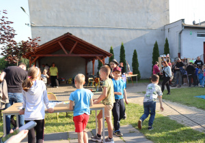 Na zdjęciu dzieci i dorośli uczestniczą w szkolnym pikniku. Na pierwszym planie kilkoro dzieci stoi przy stole, a w tle widać drewnianą wiatę, pod którą znajdują się osoby obsługujące strefę gastronomiczną. Po prawej stronie odbywa się loteria fantowa – uczestnicy stoją w kolejce przed półkami z nagrodami. Na placu panuje słoneczna pogoda i radosna, rodzinna atmosfera.