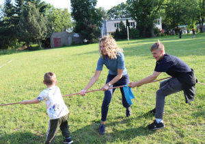 Na zdjęciu troje uczestników pikniku – mały chłopiec, nastolatek i dorosła kobieta – bierze udział w zabawie w przeciąganie liny na trawiastym boisku. Wszyscy są zaangażowani, uśmiechnięci i skupieni na rywalizacji. Kobieta znajduje się pośrodku i trzyma linę z niebieską wstążką oznaczającą środek. W tle widać przestrzeń boiska, drzewa i kilka budynków. Scena przedstawia integracyjną aktywność fizyczną podczas pikniku rodzinnego.