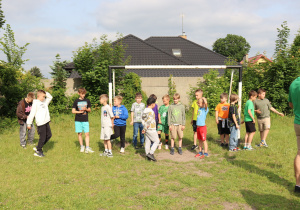 Na zdjęciu kilkunastu chłopców stoi w pobliżu bramki piłkarskiej na trawiastym boisku. Część z nich rozmawia ze sobą, niektórzy patrzą w stronę aparatu, inni są w ruchu. W tle widoczny jest wysoki płot porośnięty roślinnością oraz budynki mieszkalne z ciemnym dachem. Po prawej stronie widać fragment postaci osoby dorosłej w zielonej koszulce. Scena przedstawia przygotowania do gry w piłkę nożną w ramach pikniku lub wydarzenia sportowego.