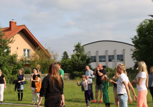 Zdjęcie przedstawia grupę osób – dorosłych i dzieci – stojących na trawie, patrzących w jednym kierunku poza kadr. W tle znajduje się biały budynek sali gimnastycznej oraz dom jednorodzinny z czerwonym dachem. Niektórzy uczestnicy trzymają ręce nad oczami, jakby osłaniali się przed słońcem. Widoczna jest swobodna atmosfera wydarzenia plenerowego, prawdopodobnie pikniku szkolnego.