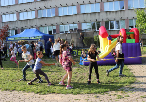 Grupa dzieci przeciąga linę podczas zabawy na świeżym powietrzu. Na drugim planie widać kolorową dmuchaną zjeżdżalnię, trampolinę oraz niebieski namiot z napisem „Park Trampolin Air Jump”. W tle stoi wielu uczestników pikniku szkolnego, w tym dzieci i dorośli. Całość odbywa się na trawniku przed budynkiem szkoły.