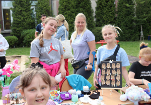 Kilka dziewczynek stoi przy stole z rękodziełem na świeżym powietrzu. Na stole rozłożone są kolorowe bransoletki, kolczyki, świeczki, maskotki i kwiaty z włóczki. Jedna z dziewczynek trzyma szydełkowy wyrób, inna siedzi i zapisuje coś na kartce. W tle widać drzewa, plac zabaw i innych uczestników pikniku.