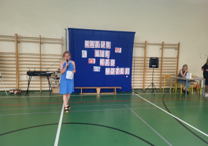 Szkolna sala gimnastyczna. Na pierwszym planie kobieta w niebieskiej sukience mówi do mikrofonu i trzyma kartkę z notatkami. Za nią znajduje się niebieski parawan z napisem „Ratuj się kto może!” oraz symbolami związanymi z pierwszą pomocą (m.in. serce, krzyż, defibrylator). Po prawej stronie przy stole siedzi nauczycielka, a obok niej stoi kolejna osoba. W tle widać drabinki gimnastyczne, sprzęt nagłaśniający i klawiaturę elektroniczną.