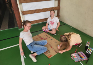 Trzy dziewczynki siedzą na zielonej podłodze sali gimnastycznej przy ścianie, tworząc plakat na dużym kartonie. Jedna z nich rysuje markerem, druga uśmiecha się do obiektywu, a trzecia odpoczywa, trzymając pisak. Obok nich leżą kolorowe flamastry i piórnik. Na plakacie widoczne są symbole związane z pierwszą pomocą, m.in. serce, krzyż i wykres EKG.