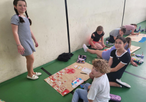 Grupa dzieci pracuje nad plakatami edukacyjnymi wzdłuż ściany sali gimnastycznej. Na pierwszym planie troje uczniów tworzy plakat z hasłem „Udzielanie pierwszej pomocy może uratować życie”, który zawiera czerwony krzyż, serce i rysunki apteczek. Dziewczynka w sukience w paski stoi obok pracy, pozując do zdjęcia. Pozostali uczniowie siedzą lub klęczą, rysując i malując. W tle widać inne grupy dzieci pochylone nad swoimi projektami.