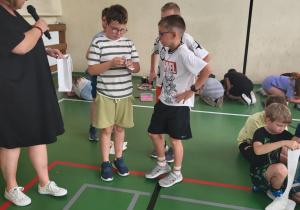 Na zdjęciu grupa chłopców stoi na zielonej podłodze sali gimnastycznej. Dwóch z nich trzyma w rękach kartki lub dyplomy, a jedna z nauczycielek w czarnej sukience i białych tenisówkach wręcza nagrodę, mówiąc do mikrofonu. W tle inne dzieci kontynuują zadania na matach. Po prawej stronie chłopiec siedzi na podłodze i rozwija rolkę papieru, jakby przygotowując się do ćwiczenia związanego z opatrywaniem.