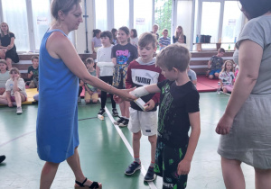 W sali gimnastycznej nauczycielka w niebieskiej sukience wręcza nagrodę chłopcu ubranemu na sportowo. W tle grupa uczniów stoi w kolejce i obserwuje wydarzenie. Widać chłopców z zabandażowanymi ramionami, co sugeruje udział w ćwiczeniach z zakresu pierwszej pomocy. Za nimi siedzą inni uczniowie na ławkach i materacach. Sala jest dobrze oświetlona naturalnym światłem wpadającym przez duże okna.