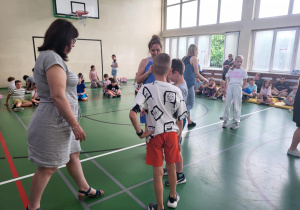 Sala gimnastyczna pełna uczniów. Na pierwszym planie dwóch chłopców stoi naprzeciw nauczycielki w niebieskiej sukience, która wręcza im nagrody. Obok stoi druga nauczycielka w jasnej sukience. W tle dzieci siedzą na podłodze, obserwując wydarzenie. Niektóre osoby rozmawiają, inne czekają w kolejce. W sali widać kosz do koszykówki i duże okna wpuszczające światło dzienne.