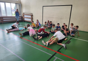 Sala gimnastyczna, na której dzieci w parach ćwiczą układanie rówieśników w pozycji bocznej ustalonej. Trzy pary uczniów wykonują ćwiczenia na podłodze, reszta dzieci siedzi z boku i przygląda się działaniom kolegów. W tle przy oknie stoi nauczycielka obserwująca zajęcia. Podłoga jest zielona, z widocznymi liniami boiska i czerwoną linią bezpieczeństwa.