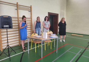 Cztery nauczycielki stoją przy stole ustawionym w sali gimnastycznej. Jedna z nich, ubrana w niebieską sukienkę, mówi do mikrofonu. Na stole leżą dokumenty, kartka i biały upominek. Za stołem stoją trzy inne nauczycielki, ubrane w codzienne stroje – jeansy, sukienkę i tunikę. W tle widoczne są drabinki gimnastyczne, głośnik i fragment otwartych drzwi.