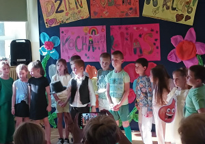 Grupa dzieci stoi na scenie w klasie szkolnej podczas występu z okazji „Dnia Rodziny”. Dzieci są przebrane w różnorodne stroje – niektóre eleganckie, inne nawiązujące do tematyki przyrody lub postaci bajkowych. W tle widoczne są kolorowe plakaty z napisami „Dzień Rodziny w kl. 1B” i „Kochamy Was” oraz duże, dekoracyjne kwiaty. W pierwszym rzędzie widać publiczność – dorośli trzymający telefony i nagrywający występ.