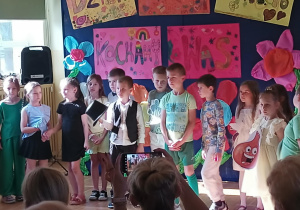 Grupa dzieci stoi na scenie szkolnej, biorąc udział w przedstawieniu z okazji Dnia Rodziny. Dzieci ubrane są w kolorowe stroje, niektóre z nich mają przebrania lub elementy kostiumów, np. rysunkową cebulę zawieszoną na szyi. W tle zawieszone są duże, ręcznie wykonane plakaty z napisami: „Dzień Rodziny w kl. 1B” i „Kochamy Was”, udekorowane kwiatami, sercami i rysunkami. Przed sceną siedzą dorośli widzowie, z których część fotografuje występ telefonami.