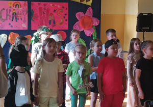 Grupa uczniów klasy 1B stoi na scenie podczas występu z okazji Dnia Rodziny. Dzieci mają na sobie kolorowe kostiumy – niektóre przebrane są za zwierzęta lub rośliny, inne ubrane w jednolite stroje tematyczne. W tle widać barwne, ręcznie wykonane plakaty z napisami „Dzień Rodziny w kl. 1B” i „Kochamy Was”, udekorowane rysunkami, sercami i dużymi kwiatami z bibuły. Po prawej stronie widać rękę dorosłej osoby nagrywającej występ telefonem.