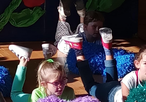 Dzieci leżą na brzuchu na podłodze sali, unosząc nogi w górę i trzymając kolorowe pompony – fioletowe, niebieskie i zielone. Dziewczynka na pierwszym planie w jasnozielonej bluzce i różowych okularach patrzy w bok, skupiona na choreografii. W tle widać dekoracje z dużych kwiatów z bibuły na ciemnoniebieskim tle. Zdjęcie wykonane podczas występu z okazji Dnia Rodziny w klasie 1B.