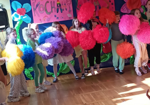 Grupa dzieci z klasy 1B wykonuje układ taneczny z dużymi, kolorowymi pomponami – w kolorach żółtym, niebieskim, fioletowym, czerwonym, różowym i pomarańczowym. Dzieci stoją na scenie przed barwną dekoracją z plakatami zawierającymi napisy „Dzień Rodziny w kl. 1B” oraz „Kochamy Was”, ozdobionymi rysunkami, sercami, kwiatami i tęczą. W tle widać również dekoracyjne kwiaty z bibuły.