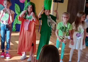 Sześć dziewczynek stoi na scenie w przebraniach symbolizujących warzywa. Każda z nich ma na ubraniu przypiętą ilustrację z uśmiechniętym warzywem, np. pomidorem, marchewką, pietruszką, szczypiorkiem i burakiem. Dzieci mają kolorowe stroje – w odcieniach zieleni, czerwieni, pomarańczu i bieli – oraz elementy dekoracyjne, np. peruki z liści czy korony z trawy. W tle widoczne są dekoracyjne kwiaty i ciemnoniebieska kotara.