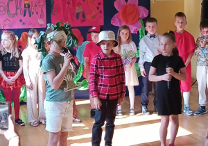 Troje dzieci stoi na środku sceny i mówi do mikrofonów – chłopiec w zielonej koszulce z opaską z liści, chłopiec w czerwonej czapce i kraciaszej koszuli oraz dziewczynka w czarnej sukience. Za nimi grupa uczniów przebranych w kolorowe stroje i warzywne przebrania. Tło zdobią ręcznie wykonane plakaty z napisami „Dzień Rodziny w kl. 1B” i „Kochamy Was” oraz papierowe kwiaty. W lewym dolnym rogu widać ręce dorosłych nagrywających wydarzenie telefonami.