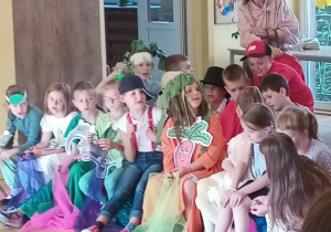 Grupa dzieci siedzi w rzędzie na krzesłach lub materacach, odpoczywając po występie szkolnym. Wiele z nich ubranych jest w kolorowe stroje z ilustracjami warzyw, np. marchewki, cebuli i pietruszki, niektóre mają nakrycia głowy z liści lub czapki. Dzieci trzymają także kolorowe tiule i pompony. W tle stoi uśmiechnięta nauczycielka przy stoliku. Scena rozgrywa się w jasnej sali z dużymi oknami i ozdobami na ścianach.