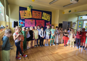 Uczniowie klasy 1B stoją w półokręgu na scenie udekorowanej plakatami z napisami „Dzień Rodziny w kl. 1B” i „Kochamy Was”. Dzieci ubrane są w różnorodne stroje – eleganckie, bajkowe i tematyczne, w tym przebrania warzyw (np. marchewka, cebula). Tło zdobią barwne kwiaty z bibuły i rysunki. W sali widoczne są dekoracje wiosenne, papierowe żaby i słoneczniki w oknach, a z sufitu zwisają ozdoby. Uczniowie pozują do wspólnego zdjęcia po zakończeniu uroczystości.