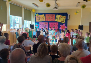 Zdjęcie wykonane podczas uroczystości szkolnej z okazji Dnia Rodziny. Na scenie przed kolorową dekoracją z napisami „Dzień Rodziny w kl. 1B” i „Kochamy Was” stoi grupa dzieci w różnorodnych strojach. Przed sceną zgromadzona jest licznie publiczność – rodzice i bliscy siedzący lub stojący, niektórzy nagrywają występ telefonami. Sala jest ozdobiona papierowymi kwiatami i żabkami, a na oknach i parapetach znajdują się dekoracje.