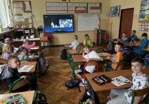 Zdjęcie przedstawia salę lekcyjną szkoły podstawowej, w której dzieci siedzą przy dwuosobowych ławkach ustawionych w rzędach. Uczniowie uśmiechają się i patrzą w stronę aparatu. Na ścianie znajduje się duży ekran, na którym wyświetlany jest czarno-biały film. W tle widać tablice, plakaty edukacyjne, komputer i meble szkolne. Na biurkach dzieci leżą przybory szkolne i kolorowanki.