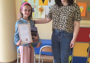 Zdjęcie przedstawia uczennicę Joannę Goszcz i nauczyciela bibliotekarza, panią Monikę Juran-Tłoczek, stojące razem w szkolnym korytarzu. Joanna trzyma w rękach dyplom oraz książkę, uśmiecha się, ma na sobie różowe spodnie, dżinsową kurtkę, różową opaskę i okulary. Obok niej stoi pani Monika w okularach, bluzce w panterkę i dżinsach, z ręką opartą na ramieniu uczennicy. W tle widoczna jest kolorowa tablica dekoracyjna z napisem „WAKACJE” oraz pracami plastycznymi przedstawiającymi lody.