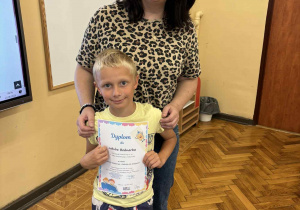 Zdjęcie przedstawia ucznia Jakuba Bednarka stojącego obok nauczyciela bibliotekarza, pani Moniki Juran-Tłoczek, w klasie szkolnej. Jakub trzyma w rękach kolorowy dyplom i uśmiecha się. Ma na sobie jasnożółtą koszulkę i krótkie spodenki z nadrukiem nosorożców. Pani Monika, ubrana w bluzkę w panterkę i dżinsy, obejmuje go delikatnie ramieniem i również się uśmiecha. W tle widoczne są: tablica, mapa Europy, drewniana podłoga i fragment ekranu multimedialnego.