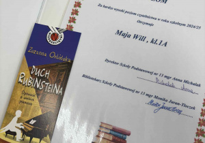 Zdjęcie przedstawia otwartą książkę z włożonym dyplomem dla uczennicy Mai Will z klasy 1A za bardzo wysoki poziom czytelnictwa w roku szkolnym 2024/2025. Dyplom opatrzony jest graficzną ramką z motywem kwiatowym, podpisany przez dyrektor szkoły mgr Annę Michalak oraz bibliotekarza szkoły Monikę Juran-Tłoczek. W lewym dolnym rogu widnieje ilustracja ułożonych książek. Do książki dołączona jest zakładka promująca tytuł „Duch Rubinsteina” autorstwa Zuzanny Orlińskiej z grafiką fortepianu i pianistki. W tle widoczna jest sala lekcyjna i uczniowie przy ławkach.