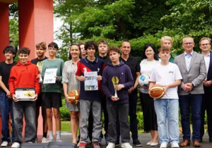 Grupowe zdjęcie przedstawiające uczniów i nauczycieli pozujących z pucharami, piłkami i dyplomami na tle czerwonego budynku i zielonych drzew. Uroczystość odbyła się 24 czerwca w ogrodzie Młodzieżowego Domu Kultury w Pabianicach z okazji rozdania nagród za współzawodnictwo sportowe szkół Powiatu Pabianickiego w roku szkolnym 2024/2025. Osoby na zdjęciu są uśmiechnięte, a część z nich trzyma trofea sportowe. Wśród obecnych znajdują się nauczyciele wychowania fizycznego – Sebastian Grzelczyk i Rafał Raźniewski.