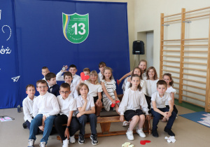 Zdjęcie grupowe przedstawia uczniów klasy trzeciej Szkoły Podstawowej nr 13 w Pabianicach oraz ich wychowawczynię. Dzieci ubrane są odświętnie – większość w białych koszulach i ciemnych spodniach lub spódniczkach. Siedzą i klęczą na ławce gimnastycznej ustawionej na sali, na tle niebieskiej dekoracji z napisem „Ruszaj w swoją podróż” oraz herbem szkoły. Na podłodze znajdują się wycięte z papieru ozdobne serca i kwiaty. W tle widoczne są drabinki gimnastyczne i głośnik. Dzieci uśmiechają się i patrzą w stronę aparatu.
