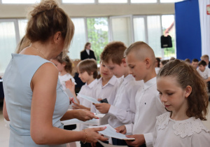 Zdjęcie przedstawia moment wręczania świadectw podczas zakończenia roku szkolnego. Po lewej stronie stoi dyrektor Szkoły Podstawowej nr 13 w Pabianicach, pani Anna Michalak, ubrana w niebieską sukienkę. Wręcza dokumenty uczniom, którzy stoją w rzędzie, ubrani w białe koszule. Dzieci z powagą i skupieniem odbierają świadectwa. W tle widać innych uczniów oraz elementy dekoracji sali, w tym fragment flagi Polski i niebiesko-białej tkaniny dekoracyjnej.