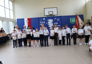 Zdjęcie przedstawia grupę uczniów klasy trzeciej stojących w rzędzie na tle dekoracji sali gimnastycznej z hasłem „Ruszaj w swoją podróż” i herbem Szkoły Podstawowej nr 13 w Pabianicach. Uczniowie ubrani są w stroje galowe i trzymają w rękach świadectwa oraz dyplomy. Za nimi stoją dyrektor szkoły, pani Anna Michalak (w niebieskiej sukience), oraz nauczycielki: Anna Wlazeł, Violetta Kocik i Agnieszka Jarzębowska. W tle widać godło Polski, sztandar szkoły i dekoracje związane z zakończeniem roku szkolnego.