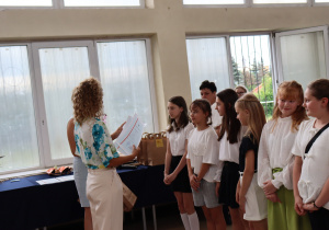 Zdjęcie przedstawia uczniów klasy 4a stojących w szeregu w sali gimnastycznej podczas uroczystości zakończenia roku szkolnego. Uczniowie ubrani są odświętnie – w białych bluzkach, koszulach, spódniczkach i spodenkach. Po lewej stronie stoi nauczycielka – wychowawczyni klasy, pani Paulina Klemżyńska, ubrana w jasne spodnie i kwiecistą bluzkę. Trzyma w rękach świadectwa, które wręcza kolejnym uczniom. W tle widać otwarte okna, papierowe torby z upominkami oraz inne osoby obserwujące wydarzenie.