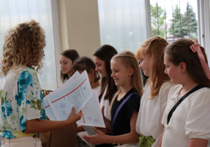 Zdjęcie przedstawia uczniów klasy 4a stojących w szeregu w sali gimnastycznej podczas uroczystości zakończenia roku szkolnego. Uczniowie ubrani są odświętnie – w białych bluzkach, koszulach, spódniczkach i spodenkach. Po lewej stronie stoi nauczycielka – wychowawczyni klasy, pani Paulina Klemżyńska, ubrana w jasne spodnie i kwiecistą bluzkę. Trzyma w rękach świadectwa, które wręcza kolejnym uczniom. W tle widać otwarte okna, papierowe torby z upominkami oraz inne osoby obserwujące wydarzenie.