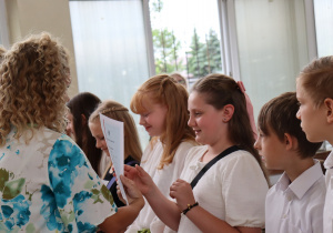 Zdjęcie przedstawia uczniów klasy 4a stojących w szeregu w sali gimnastycznej podczas uroczystości zakończenia roku szkolnego. Uczniowie ubrani są odświętnie – w białych bluzkach, koszulach, spódniczkach i spodenkach. Po lewej stronie stoi nauczycielka – wychowawczyni klasy, pani Paulina Klemżyńska, ubrana w jasne spodnie i kwiecistą bluzkę. Trzyma w rękach świadectwa, które wręcza kolejnym uczniom. W tle widać otwarte okna, papierowe torby z upominkami oraz inne osoby obserwujące wydarzenie.